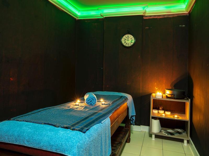 Dunkler Massageraum mit blauer Liege, Handtuch, Kerze und grüner LED-Beleuchtung an der Decke.