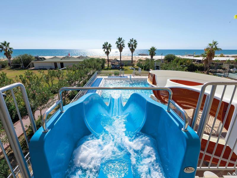 Blauer Wasserrutsche mit spritzendem Wasser, Palmen und Meer im Hintergrund an sonnigem Tag.