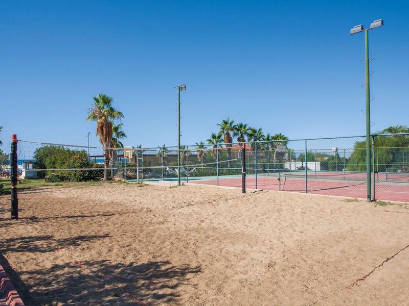 Sandplatz neben einem Tennisplatz mit rotem Belag und grünem Zaun bei klarem Himmel.