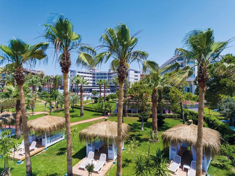 Grüner Palmenpark mit Strohdach-Liegen und modernen Gebäuden im Hintergrund bei blauem Himmel.