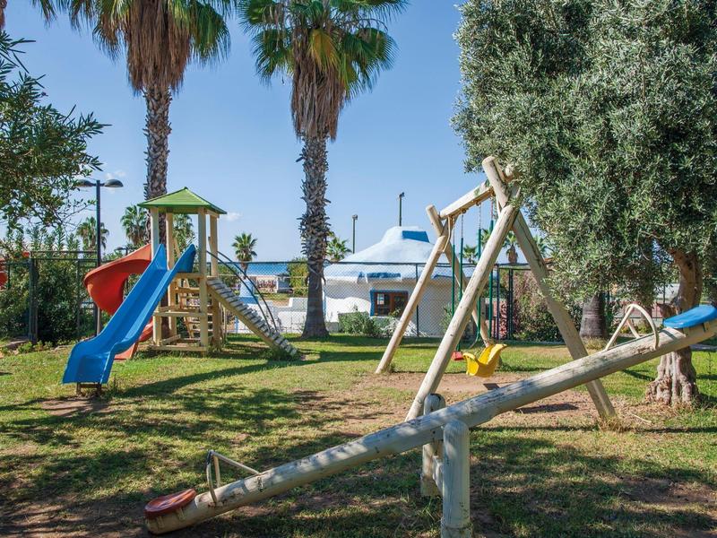 Spielplatz mit Schaukel, Rutsche, Wippe und Palmen vor blauem Himmel und Wasser.