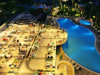 Vista notturna di una piscina di hotel illuminata con aree salotto e mare sullo sfondo.