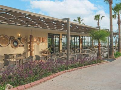 Area esterna moderna di un ristorante con tavoli, sedie e palme sotto un cielo nuvoloso.