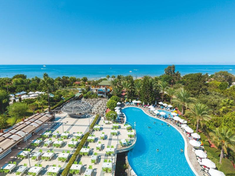 Vista di una piscina dell'hotel con ombrelloni e lettini, circondata da palme e mare sullo sfondo.