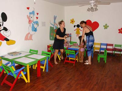 Kindergartenraum mit bunten Tischen und Stühlen, Wandbildern von Disney-Figuren und drei Personen.