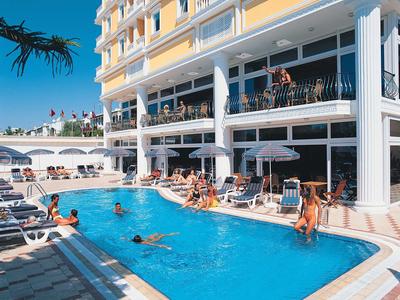 Hotelpool mit Schwimmern, Liegestühlen und Balkon, umgeben von gelbweißem Gebäude bei Sonnenschein.