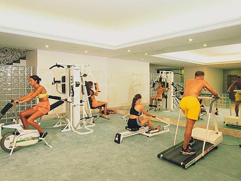 Wnętrze siłowni z trzema osobami korzystającymi z różnych urządzeń do ćwiczeń.
