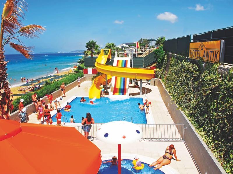 Hotelowy basen z zjeżdżalniami przy plaży, palmy i parasole, wielu gości korzysta z ciepłej pogody.