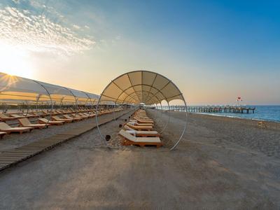 Strand mit Liegestühlen und Schirmen, dahinter Meer und Steg bei Sonnenuntergang.