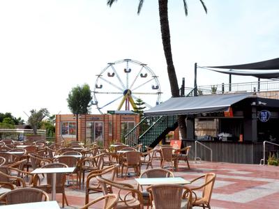 Leerer Außenbereich eines Cafés mit Tischen, Stühlen, einer Palme und einem Riesenrad im Hintergrund.