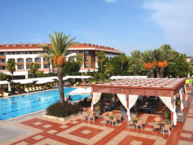 Piscina dell'hotel con lettini, padiglione e palme sotto un cielo azzurro.