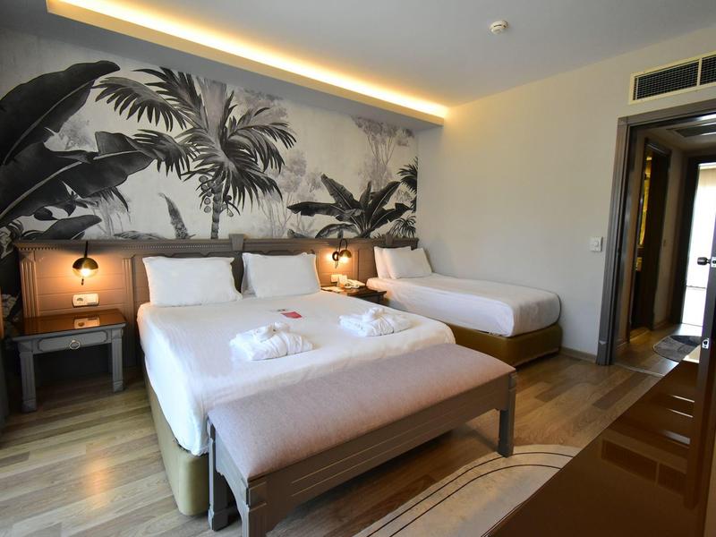 Camera d'hotel luminosa con tre letti, comodini e grande murale di piante tropicali.