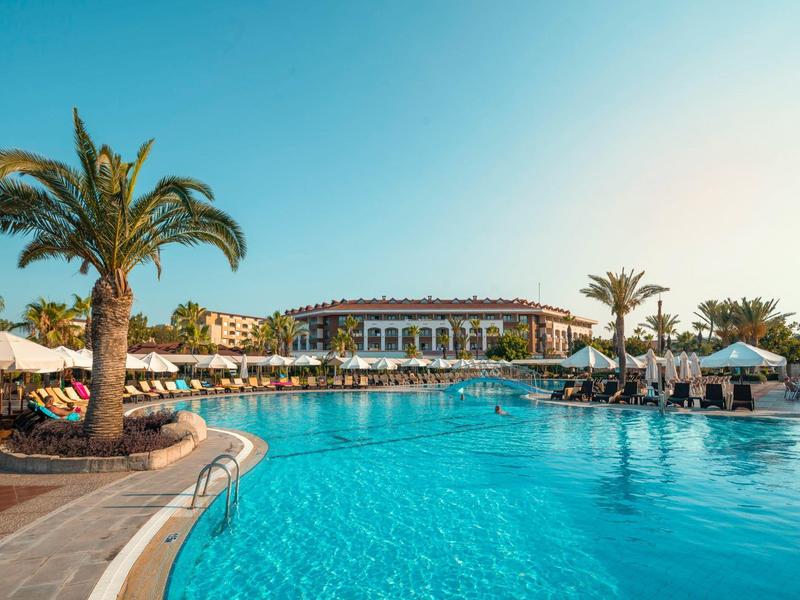 Grande piscina dell'hotel circondata da palme e lettini sotto un cielo azzurro limpido.