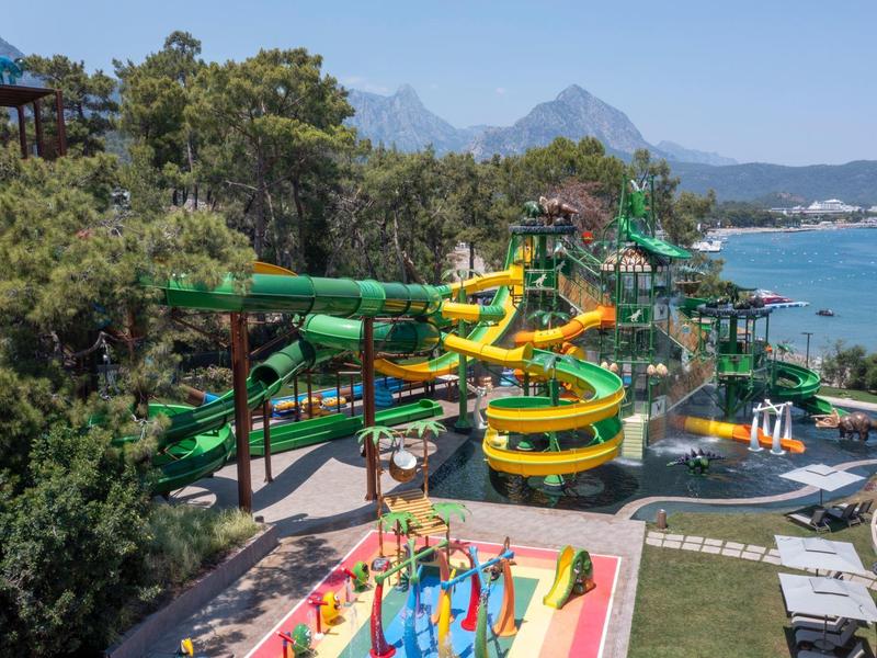 Toboganes acuáticos coloridos y una zona de juegos en un parque acuático junto al mar con montañas al fondo.
