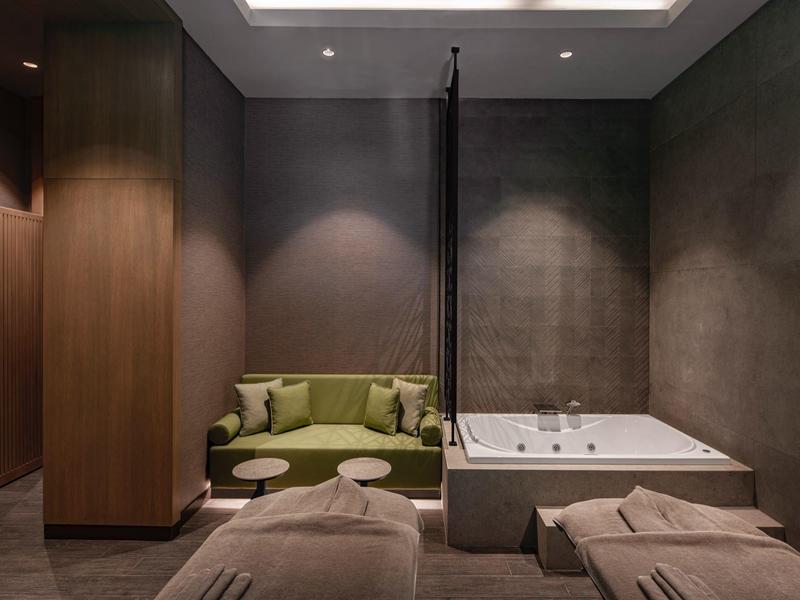 Sala de spa con dos camillas, sofá verde y bañera de hidromasaje en ambiente moderno y acogedor.