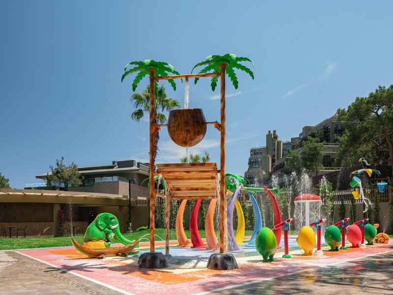 Parque acuático colorido con palmeras, chorros de agua y toboganes al aire libre bajo cielo despejado.