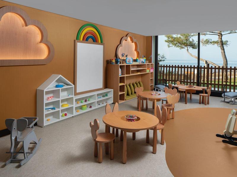 Área de juegos infantil luminosa con muebles coloridos, estanterías y vista a la terraza con árboles y mar.