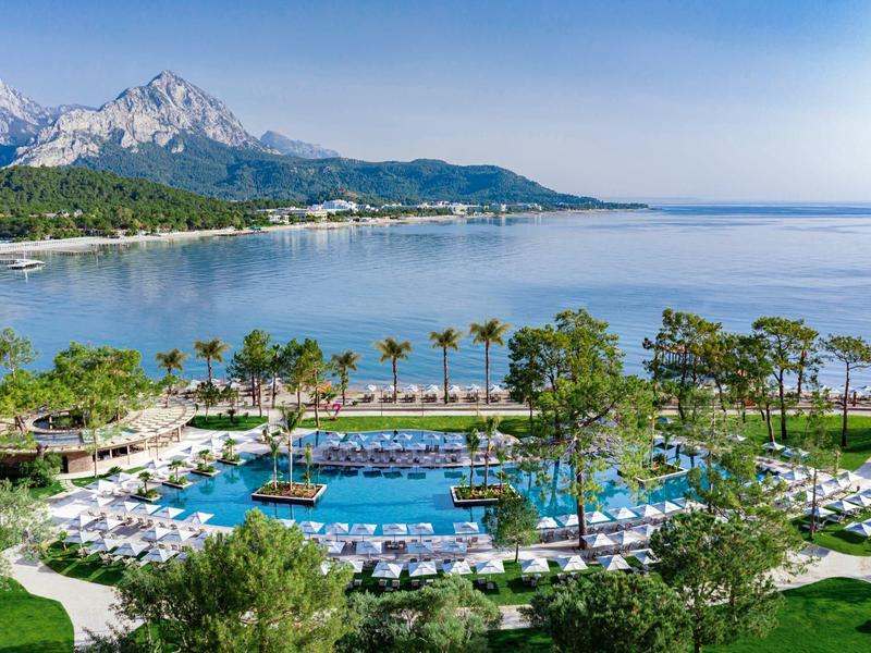 Gran piscina del hotel con tumbonas, palmeras y montañas al fondo junto al mar.