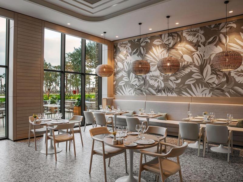 Área de restaurante de hotel moderno con mesas y sillas de madera y grandes ventanales con vista exterior.