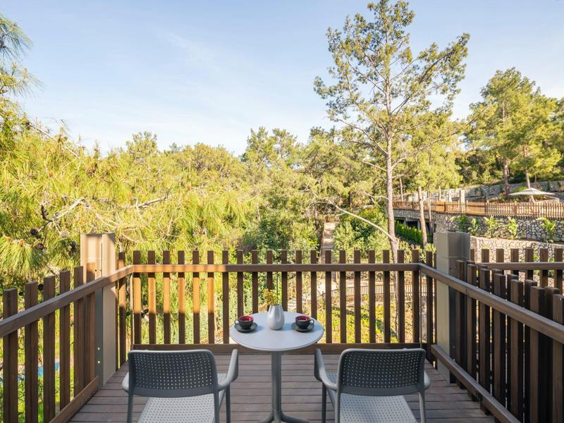 Balcón con dos sillas y mesa pequeña, rodeado de barandilla de madera y árboles.