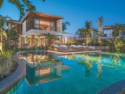 Moderner Pool mit klarem Wasser vor luxuriösen Villen und Palmen unter blauem Himmel.