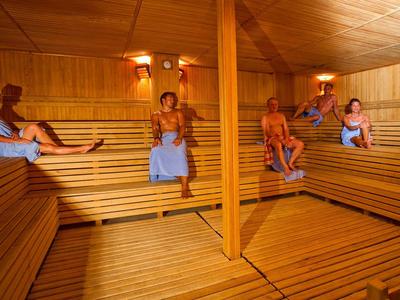 Several people sit in a wood-paneled sauna room wearing towels.