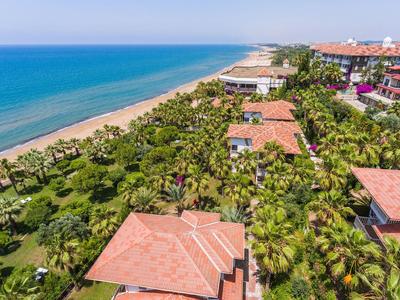 Vista de un complejo costero con playa, jardín frondoso y varios edificios con techos rojos.