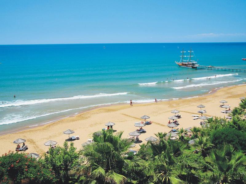 Beach with umbrellas, palm trees, and a sailing ship on the turquoise sea.