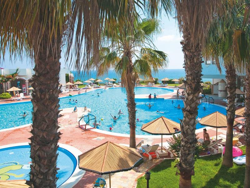 Large hotel pool with palm trees, umbrellas, and a view of the sea in the background.