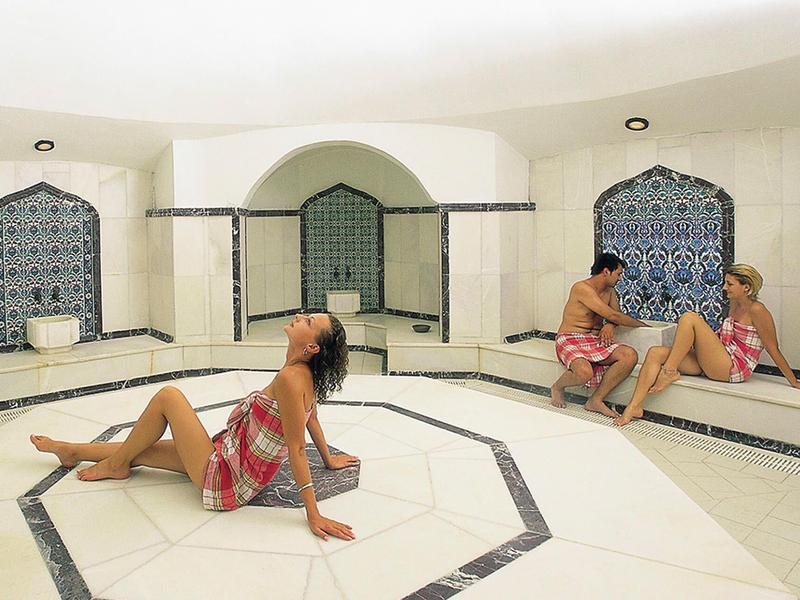 Three people relax in a bright, Arabesque-style spa with tiled benches.