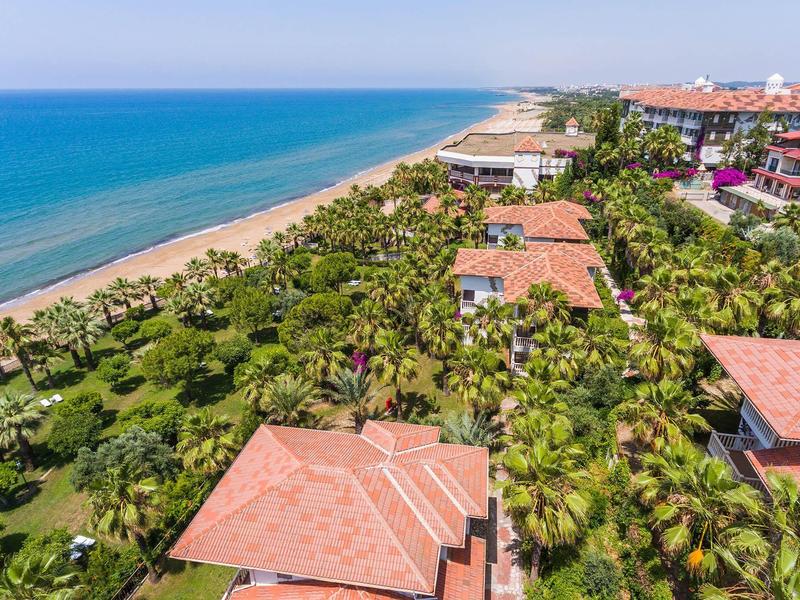 Aerial view of a hotel with red roofs by a beach lined with palm trees and blue sea.