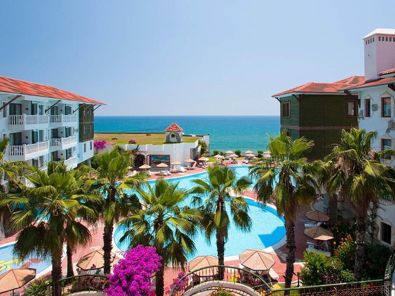 View of hotel pool with palm trees, flowering plants, and the sea in the background.