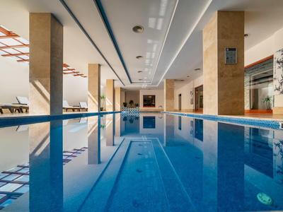 Piscina coperta con acqua blu chiara, pilastri beige e luci a soffitto in un hotel.