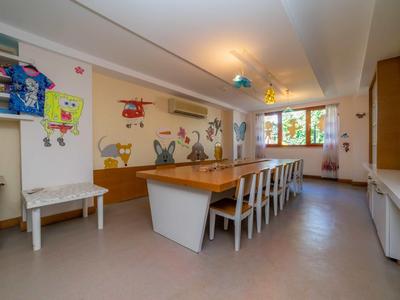 Sala giochi luminosa con lunghe tavole di legno, decorazioni murali colorate e sedie per bambini.