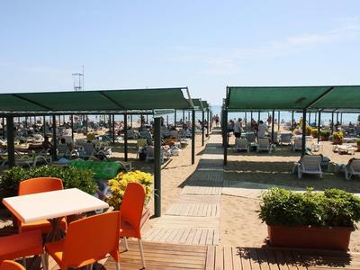 Area esterna con sedie arancioni e tavoli vicino a bancarelle coperti da tendoni verdi in una giornata di sole.