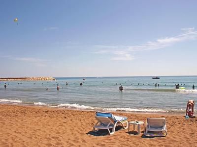 Strand mit rotem Sand, zwei blauen Liegestühlen, Tischen und Badegästen im Meer unter blauem Himmel.
