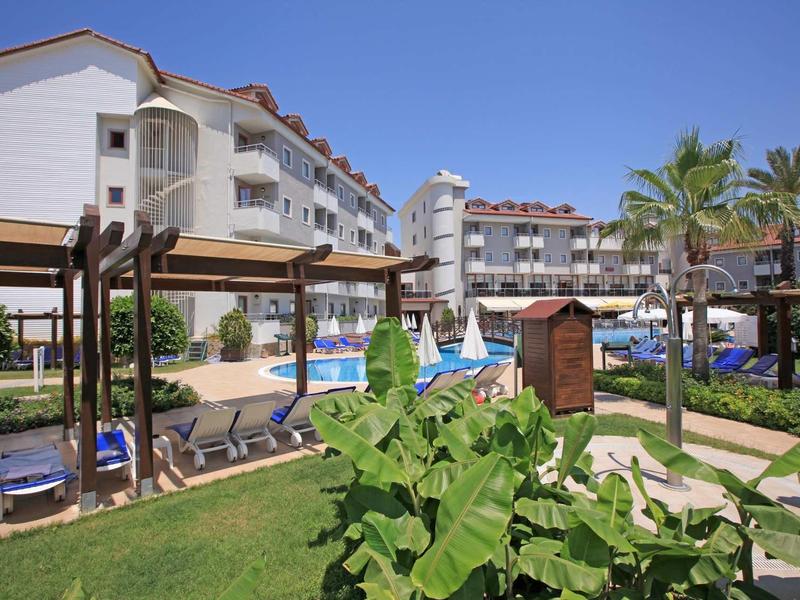 Resort con piscina, lettini, palme e edifici su più piani sotto un cielo sereno.