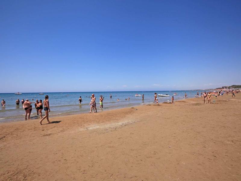 Spiaggia di sabbia con persone che camminano e nuotano sotto un cielo azzurro.