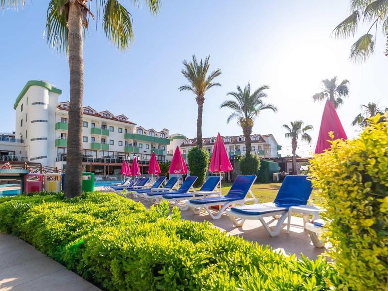 Zona piscina al sole con lettini blu, ombrelloni rosa, palme e edificio dell'hotel.
