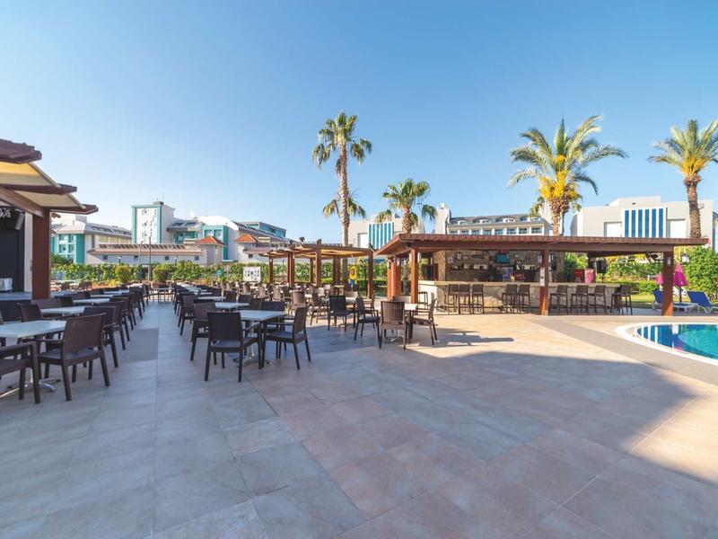 Ampio patio esterno dell'hotel con tavoli, sedie, palme e cielo azzurro.