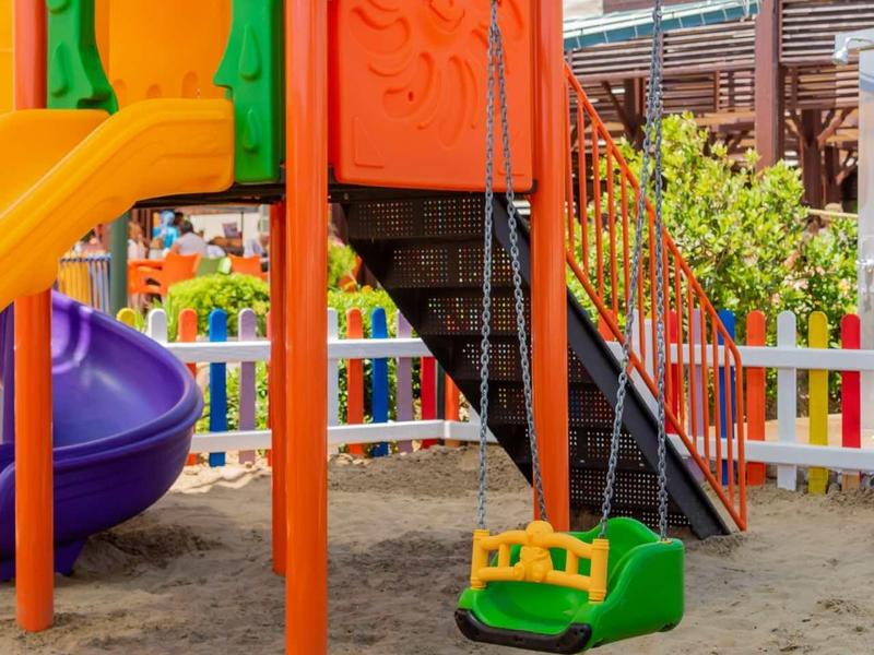 Parco giochi colorato con scivolo, scalette e altalena su sabbia vicino a recinzione bianca.