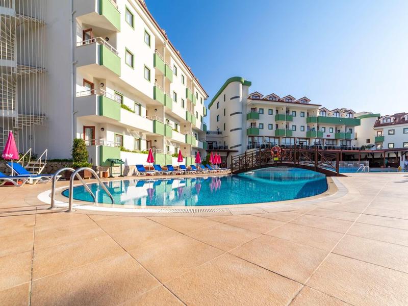 Area piscina esterna dell'hotel con lettini e ombrelloni accanto a edifici multipiano sotto un cielo blu.