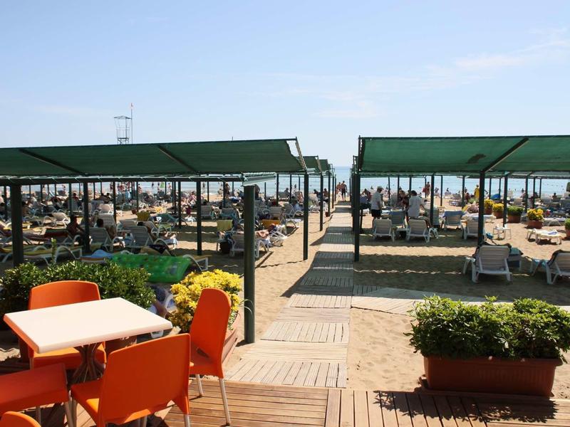 Area esterna con sedie arancioni e tavoli vicino a bancarelle coperti da tendoni verdi in una giornata di sole.