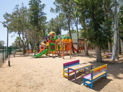 Barevné dětské hřiště se skluzavkami a lezecím rámem, obklopené stromy a pískem.