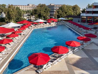 Bazén s červenými slunečníky a lehátky vedle hotelu pod modrou oblohou.