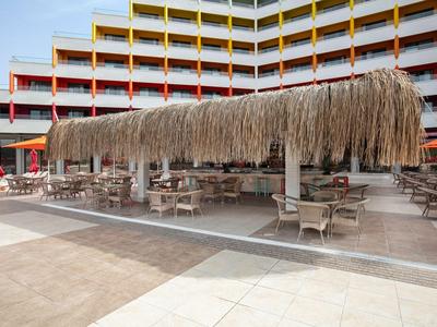 Krytá venkovní plocha s doškovou střechou a stoly před vícepatrovou hotelovou budovou.