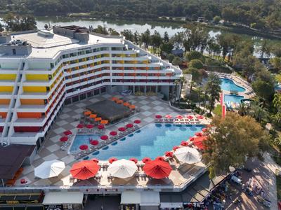 Moderní hotel s vícepodlažní budovou, protáhlým bazénem a lehátky s červenými slunečníky.