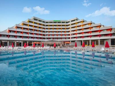 Moderní vícepodlažní hotel s velkým bazénem, červenými slunečníky a lehátky.