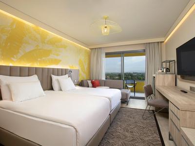 Moderní hotelový pokoj se dvěma postelemi, stolem, televizí a balkonem s výhledem.