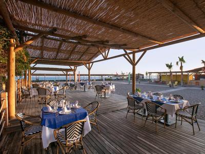 Otevřená restaurace s stolky a židlemi, výhled na moře pod dřevěnou stříškou.